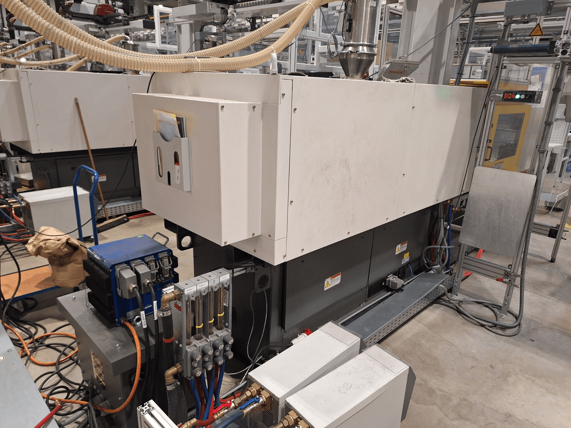 Teollisuuslaitteet tehdasympäristössä; sisältää suuren koneen, jossa on ohjauspaneeli, letkuja ja sähköliitäntöjä, sivukuva.
