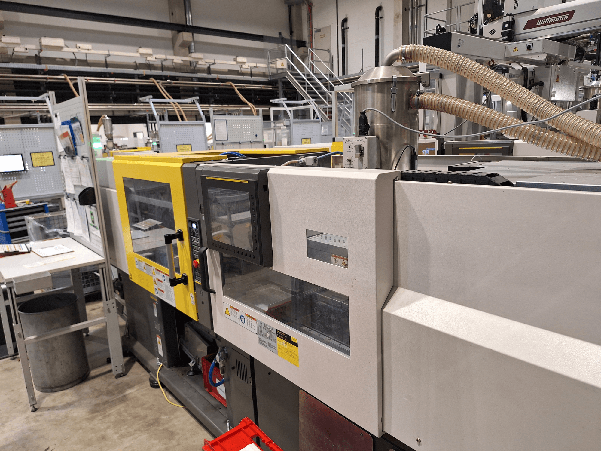 Etukuva monivärisestä ruiskuvalukoneesta, jossa on keltaisia ja harmaita osia, ohjauspaneeli ja pakojärjestelmä.