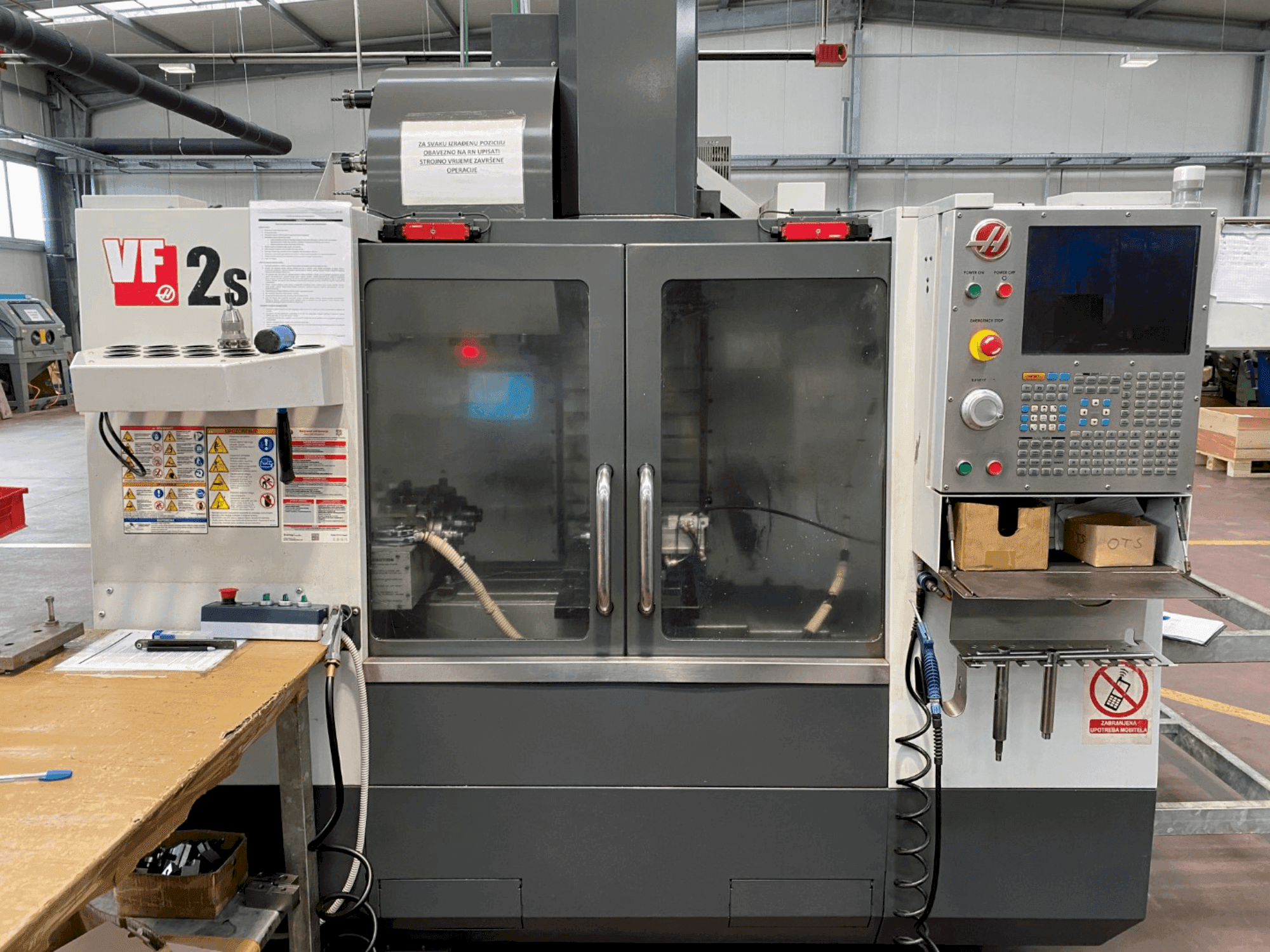 Haas VF-2SS -työstökeskus edestä, jossa on ohjauspaneeli, jäähdytysnesteletku ja sivussa olevat turvatarrat.